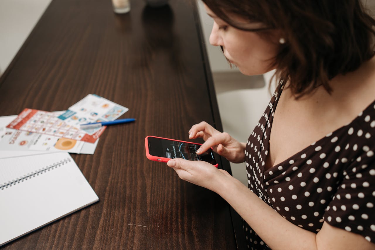 Woman in a polka dot dress using smartphone at a table indoors with documents.